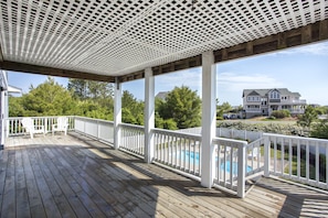A wooden deck with white railings, a pool, and a large house in the background. - Calypso: Private Pool, Hot Tub, 6 Bedrooms (Corolla)