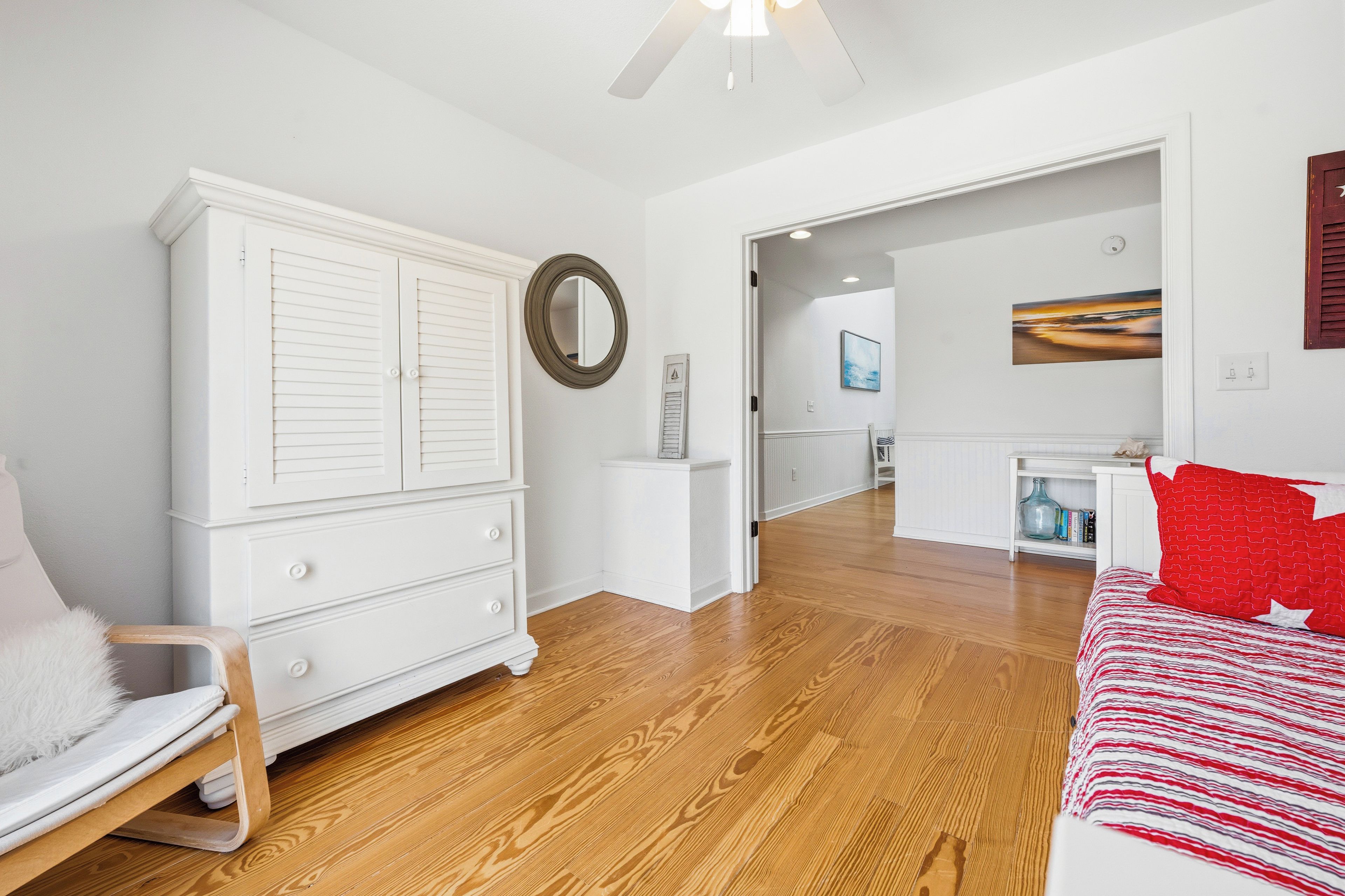 A bedroom with a wooden floor, white furniture, a bed with red pillows, a white wardrobe, a mirror, and a painting on the wall.