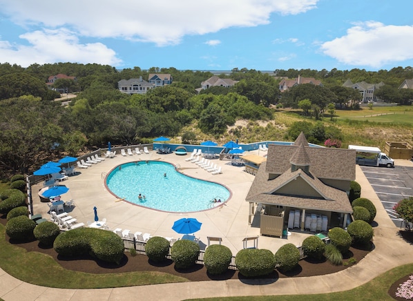 A swimming pool with lounge chairs and umbrellas, surrounded by well-maintained bushes and a building with a pitched roof. - Calypso: Private Pool, Hot Tub, 6 Bedrooms (Corolla)