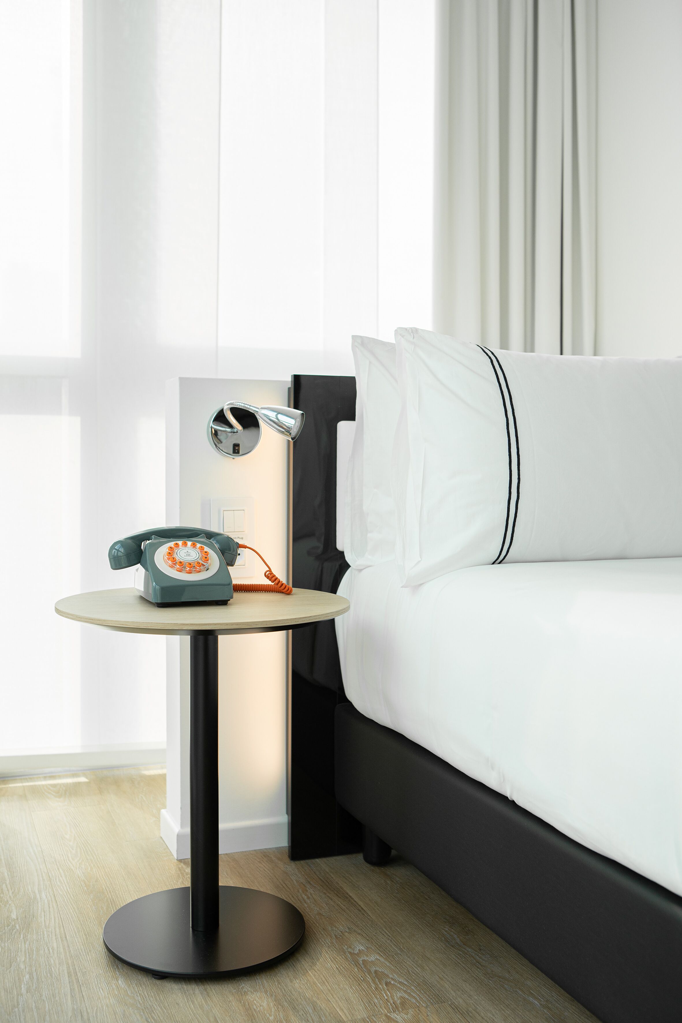 A modern hotel room with a bed, a bedside table with a rotary phone, a lamp, and a view of the outdoors through sheer curtains.