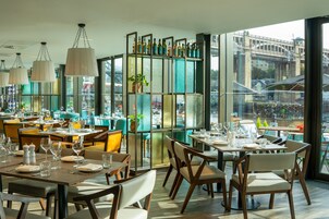 Un ristorante con diversi tavoli e sedie, ampie finestre che offrono vista su un ponte e sullo skyline cittadino, e una mensola decorativa con bottiglie.