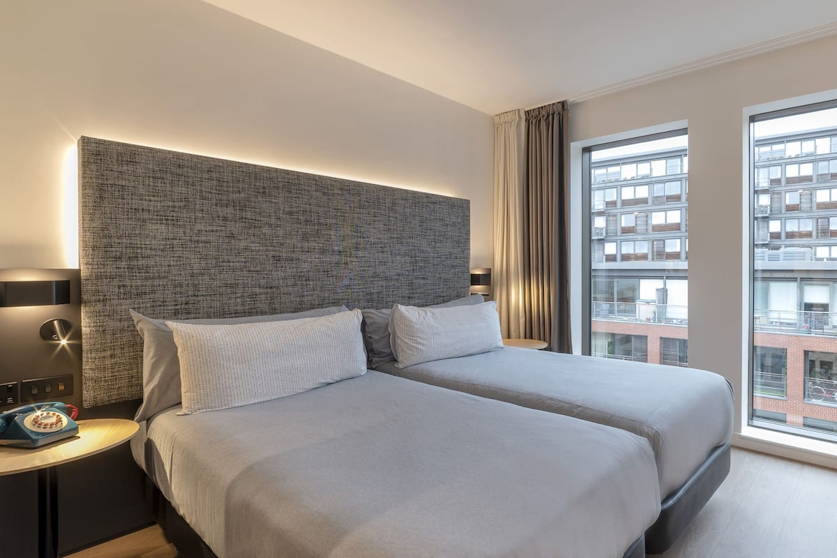 Une chambre d’hôtel moderne avec deux lits, une grande fenêtre donnant sur la ville, une table ronde et un téléphone sur la table de chevet.