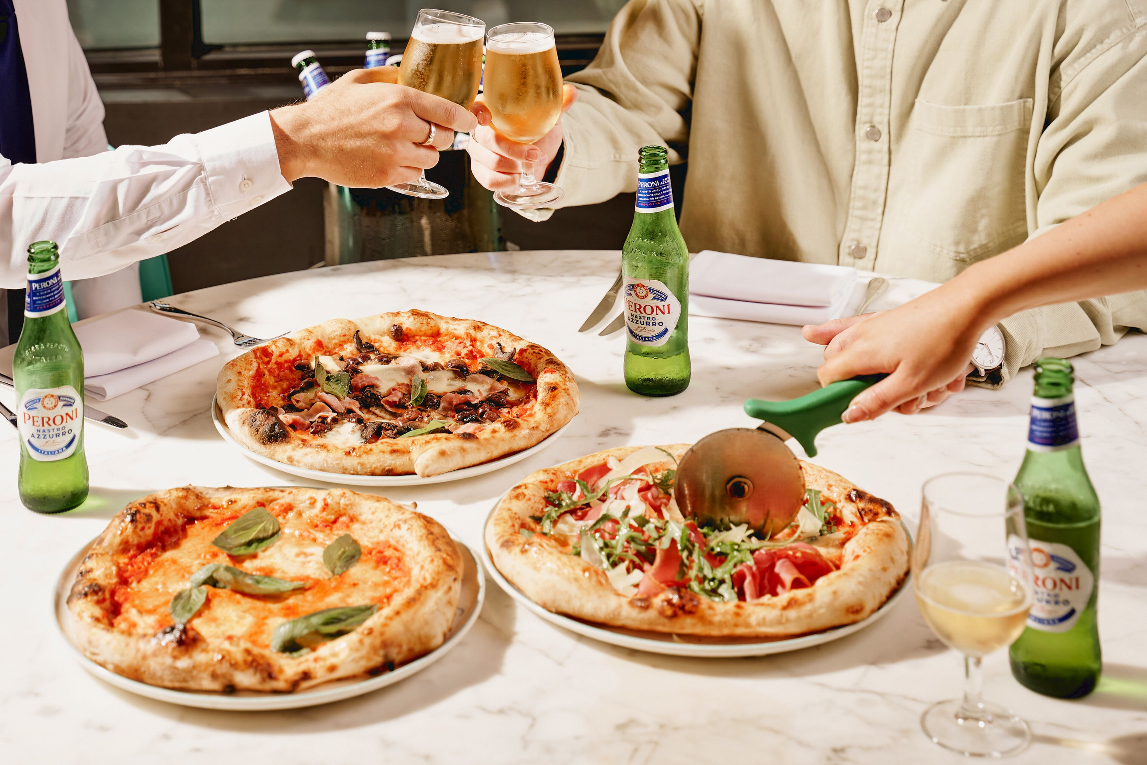 A table with three pizzas, two bottles of Peroni beer, and a pizza cutter.