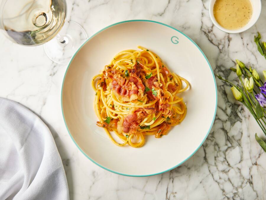 A plate of spaghetti with bacon and herbs, a glass of white wine, and a small bowl of sauce on a marble surface.
