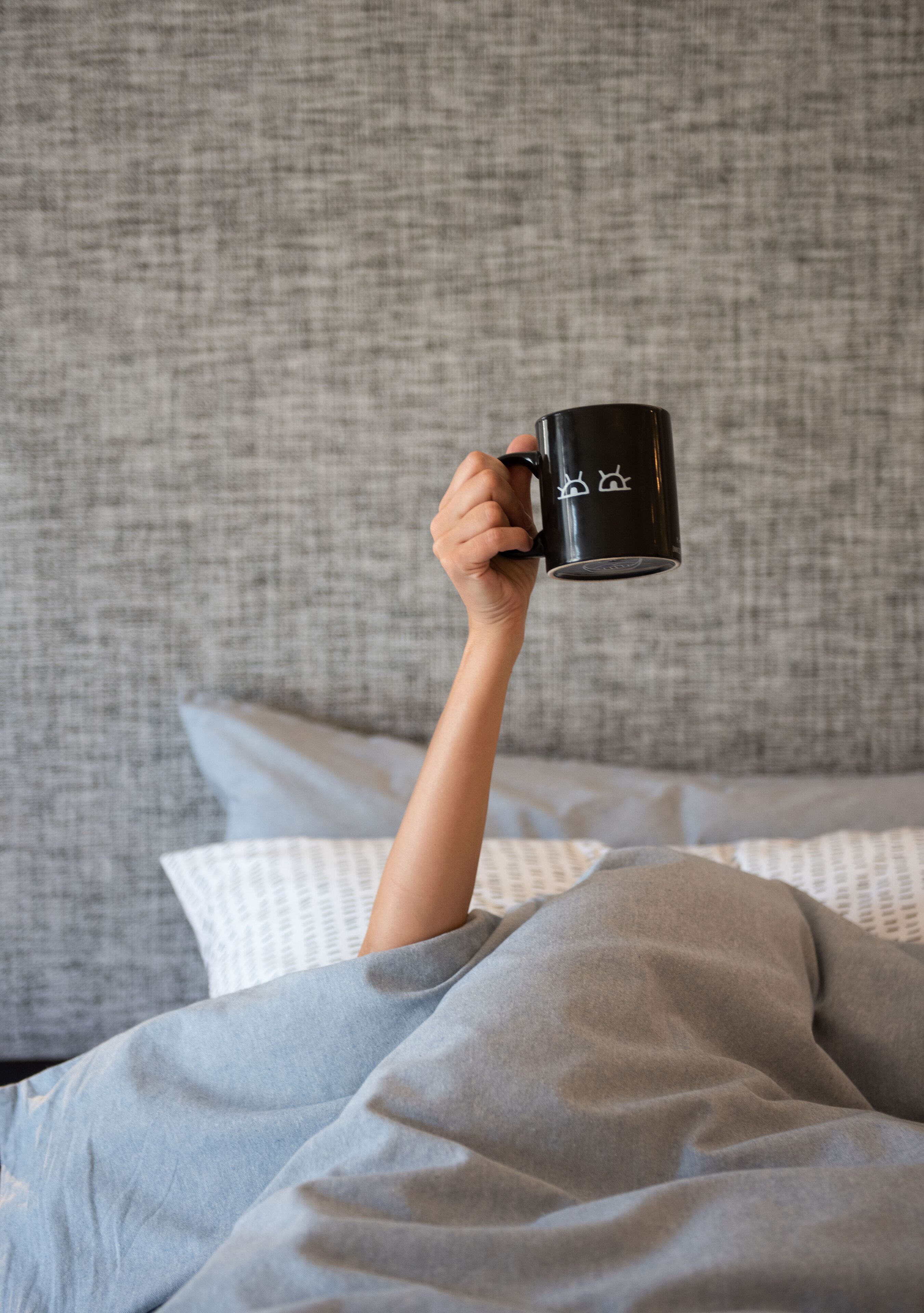 A person holding a black mug with white designs, lying in bed with grey bedding and pillows.