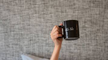 A person holding a black mug with white designs, lying in bed with grey bedding and pillows.