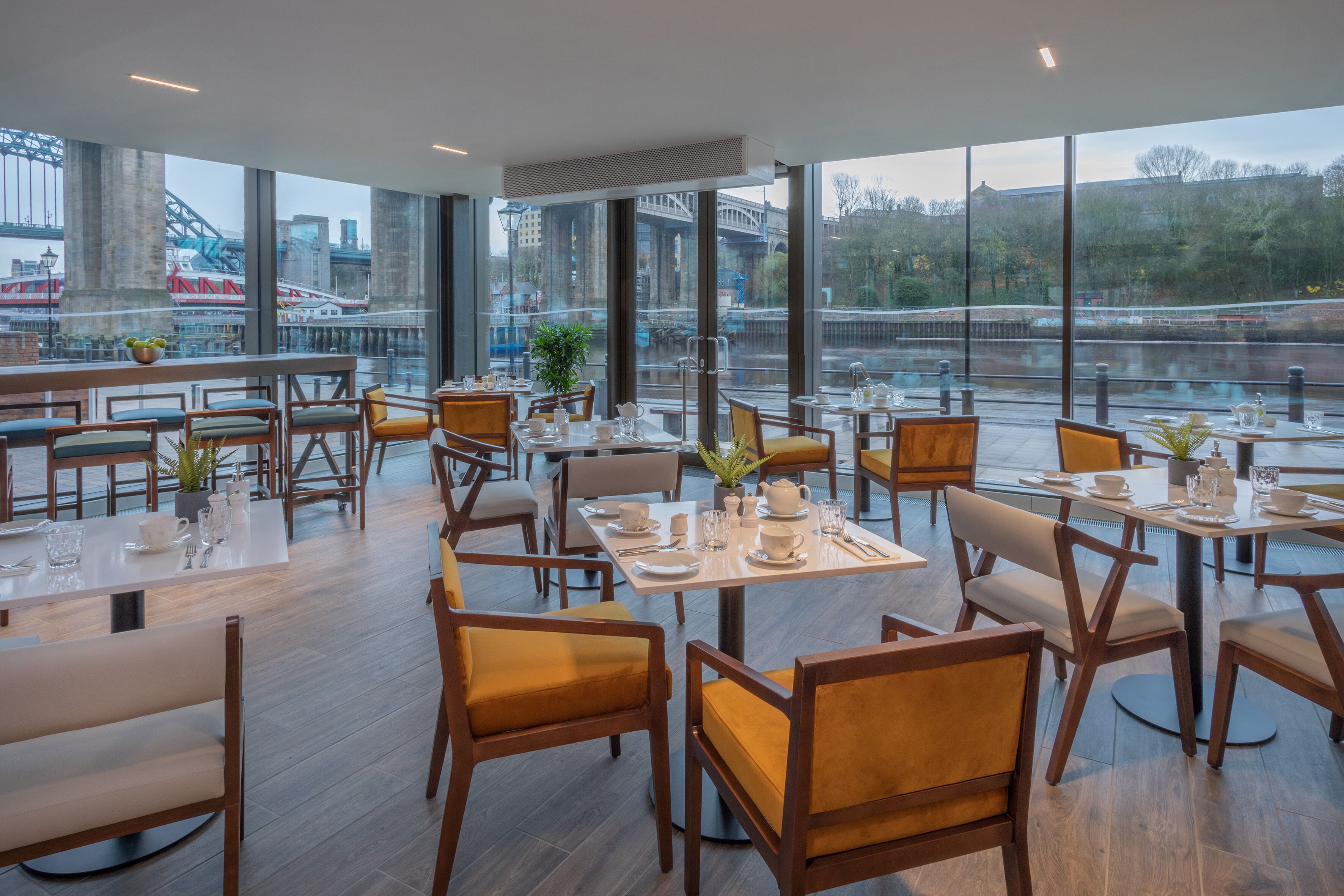 Uma sala de jantar moderna com mesas preparadas para o serviço, amplas janelas com vista para uma ponte e um rio, e um design interior limpo e contemporâneo.