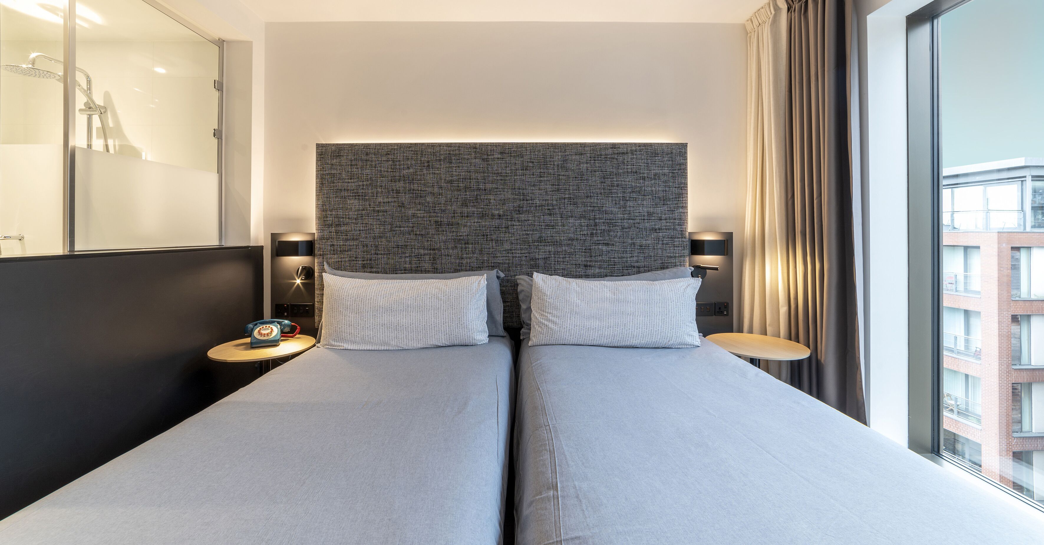 Une chambre d’hôtel moderne avec un grand lit, deux tables de chevet, une tête de lit et une vue sur un immeuble par la fenêtre.