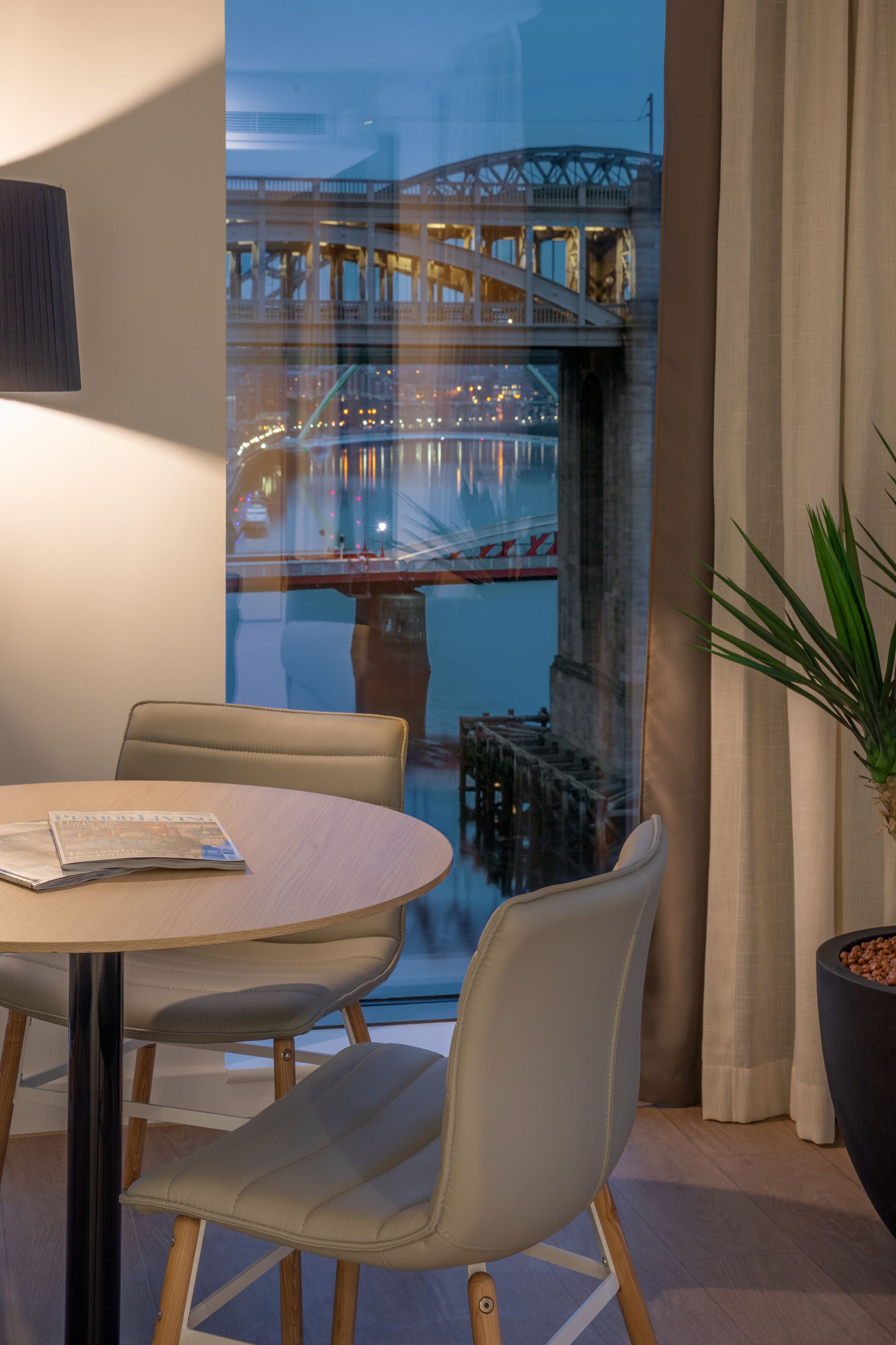 Een moderne eetruimte met een ronde tafel, twee stoelen, een potplant en uitzicht op een brug over het water.