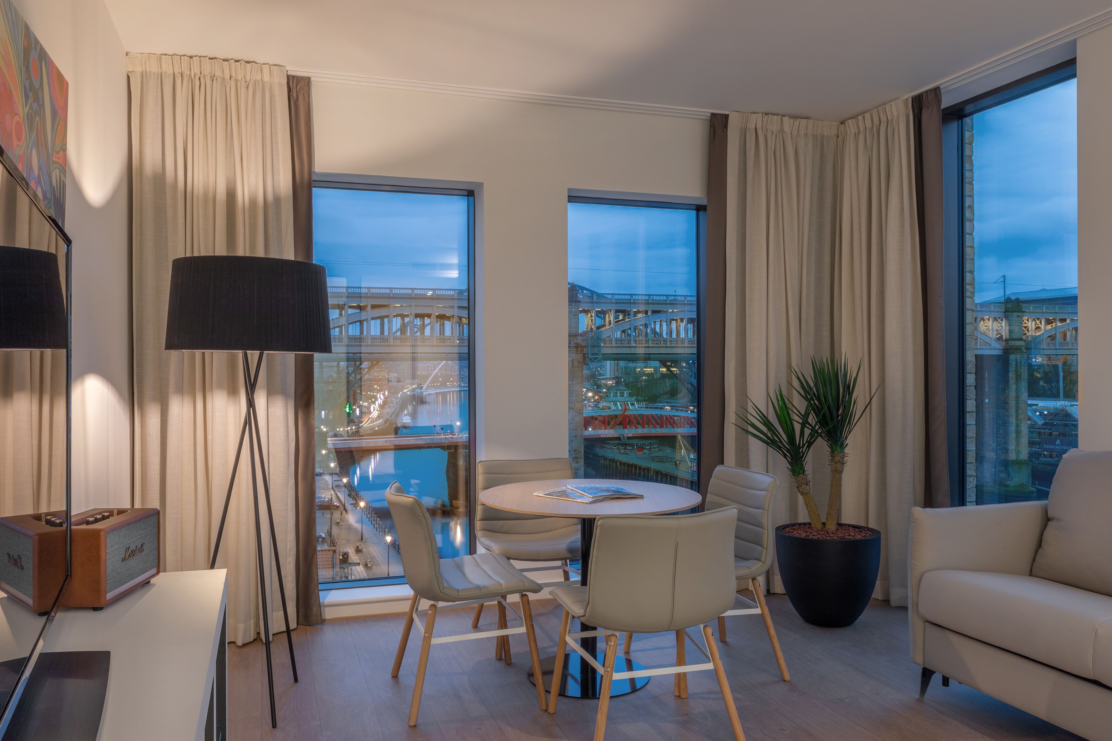 Ein modernes Hotelzimmer mit einem runden Esstisch, bequemen Sitzgelegenheiten, einer großen Pflanze und großen Fenstern mit Blick auf die Stadt.