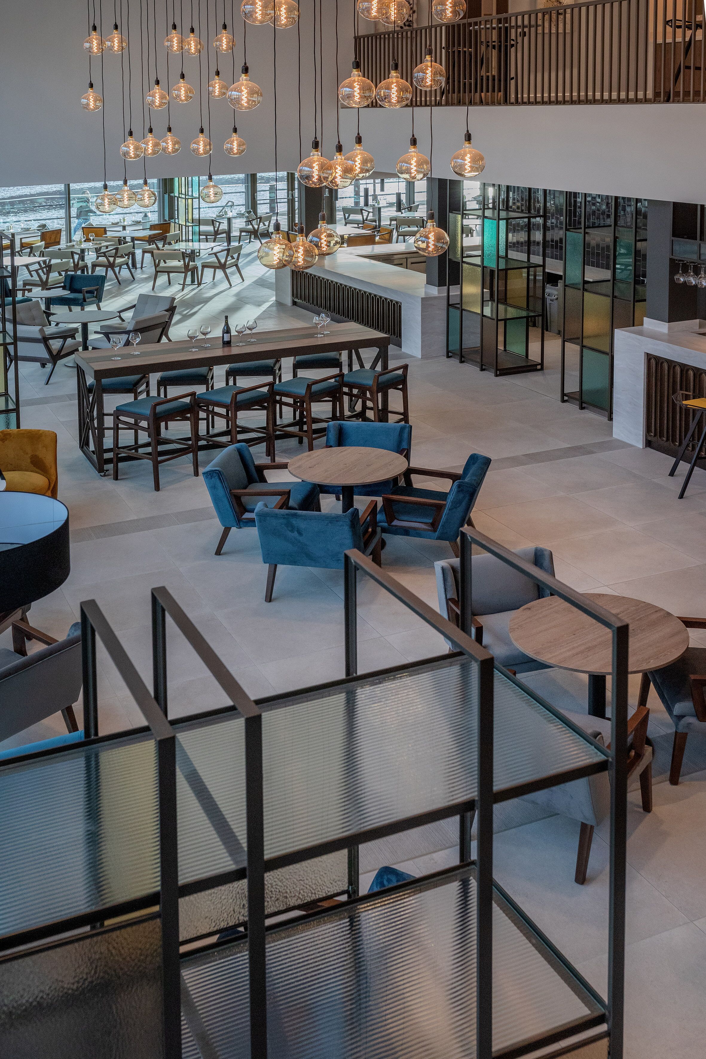 A modern interior with multiple seating areas, including round tables and sofas, and a high ceiling with hanging light fixtures.