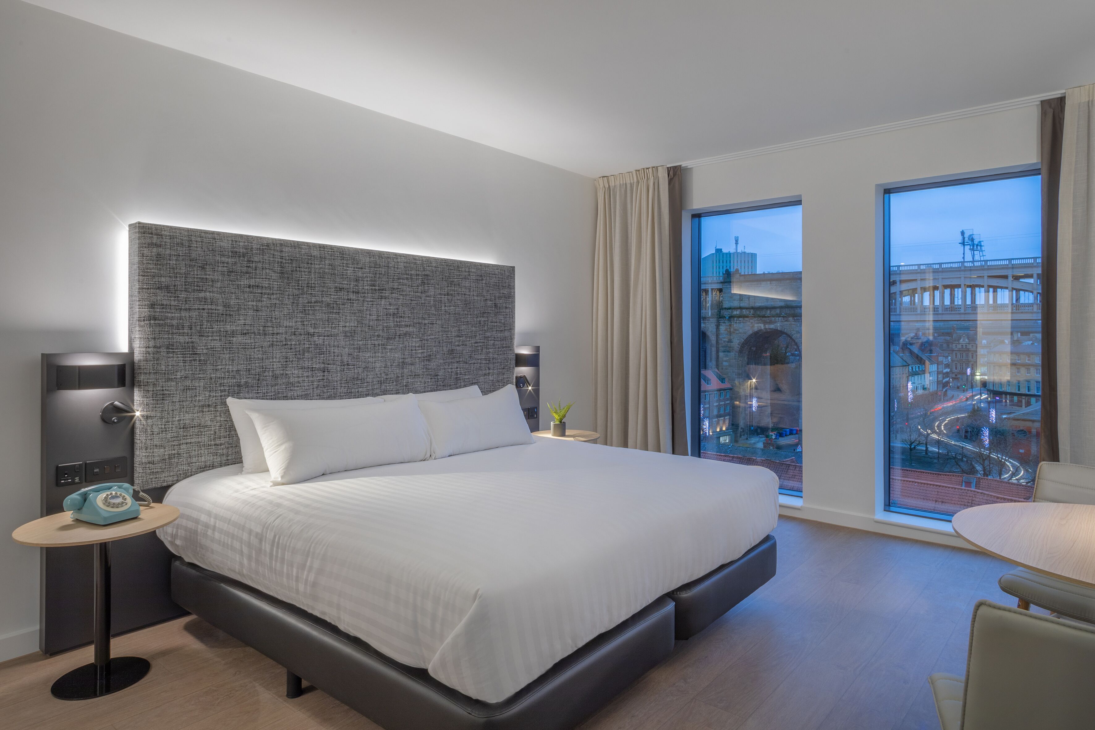 A modern hotel room with a large bed, bedside tables, a small table, and a view of a cityscape through large windows.