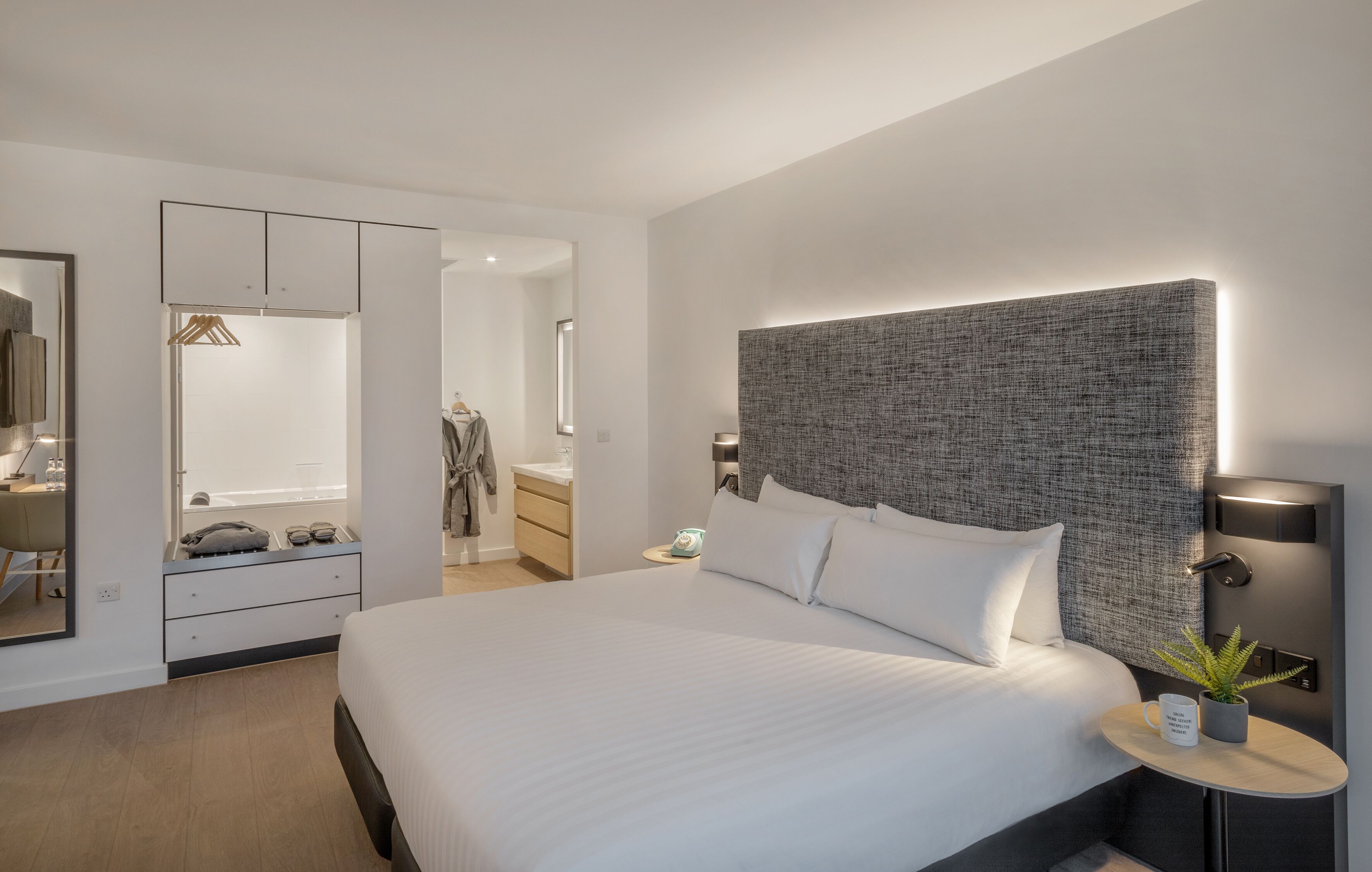 Una habitación de hotel moderna con una cama grande, un armario, una mesita de noche con lámpara y una pequeña planta en maceta.