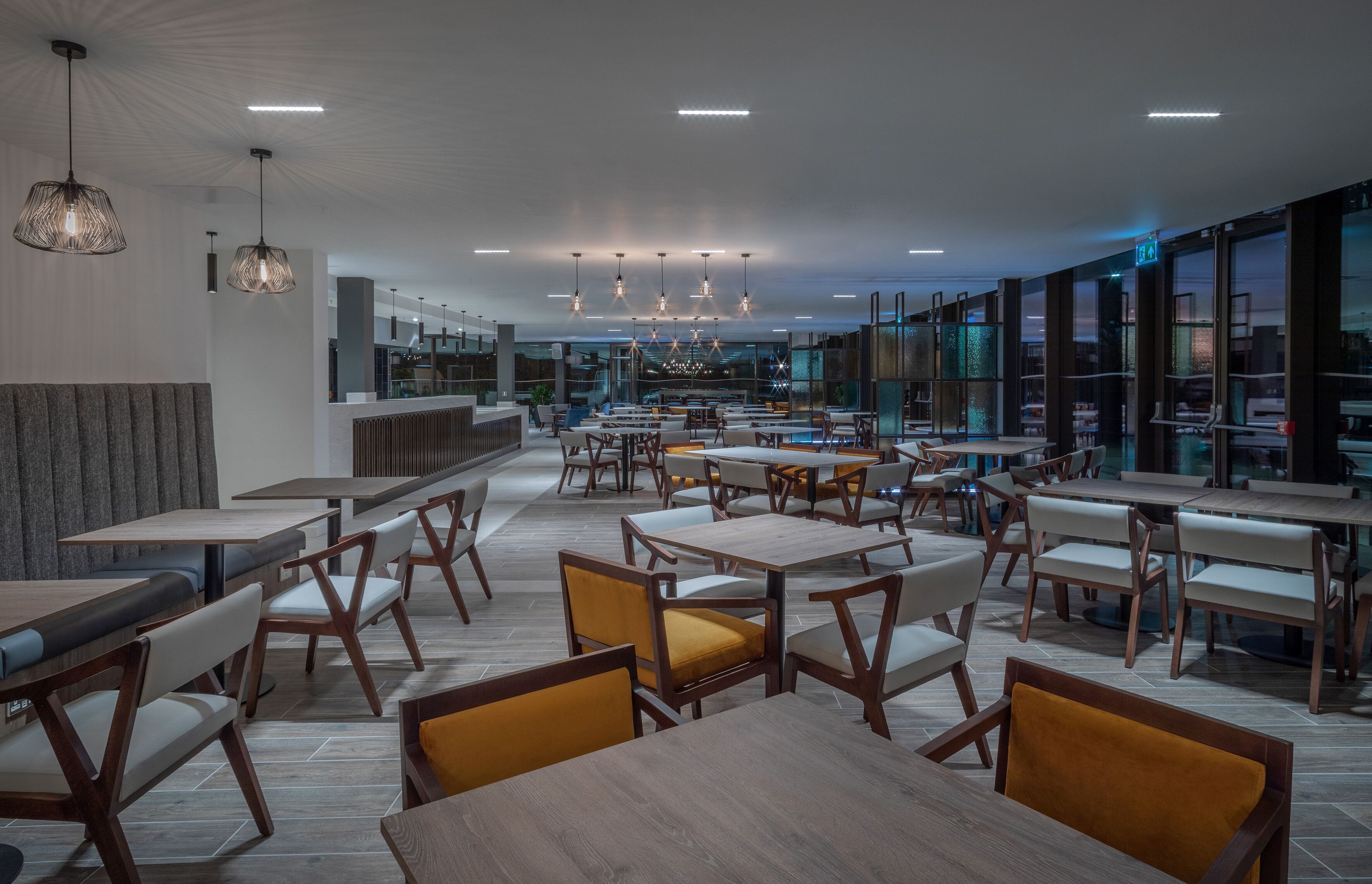 En moderne restaurant med flere spiseborde og stole, højt til loftet med hængende lamper, og store vinduer der giver udsigt over byen.
