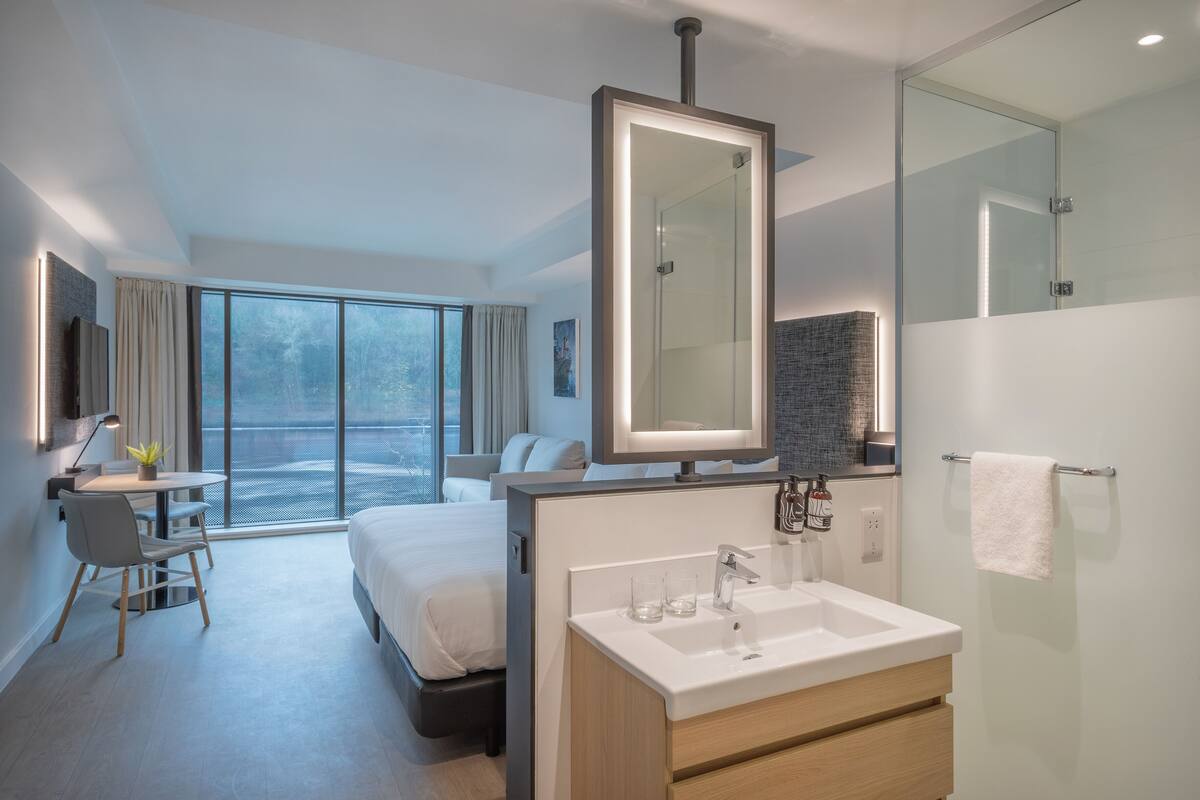 Une chambre d’hôtel moderne dotée d’un grand lit, d’un coin salon avec un bureau et une chaise, d’une salle de bain avec un grand miroir et offrant une vue sur l’extérieur.