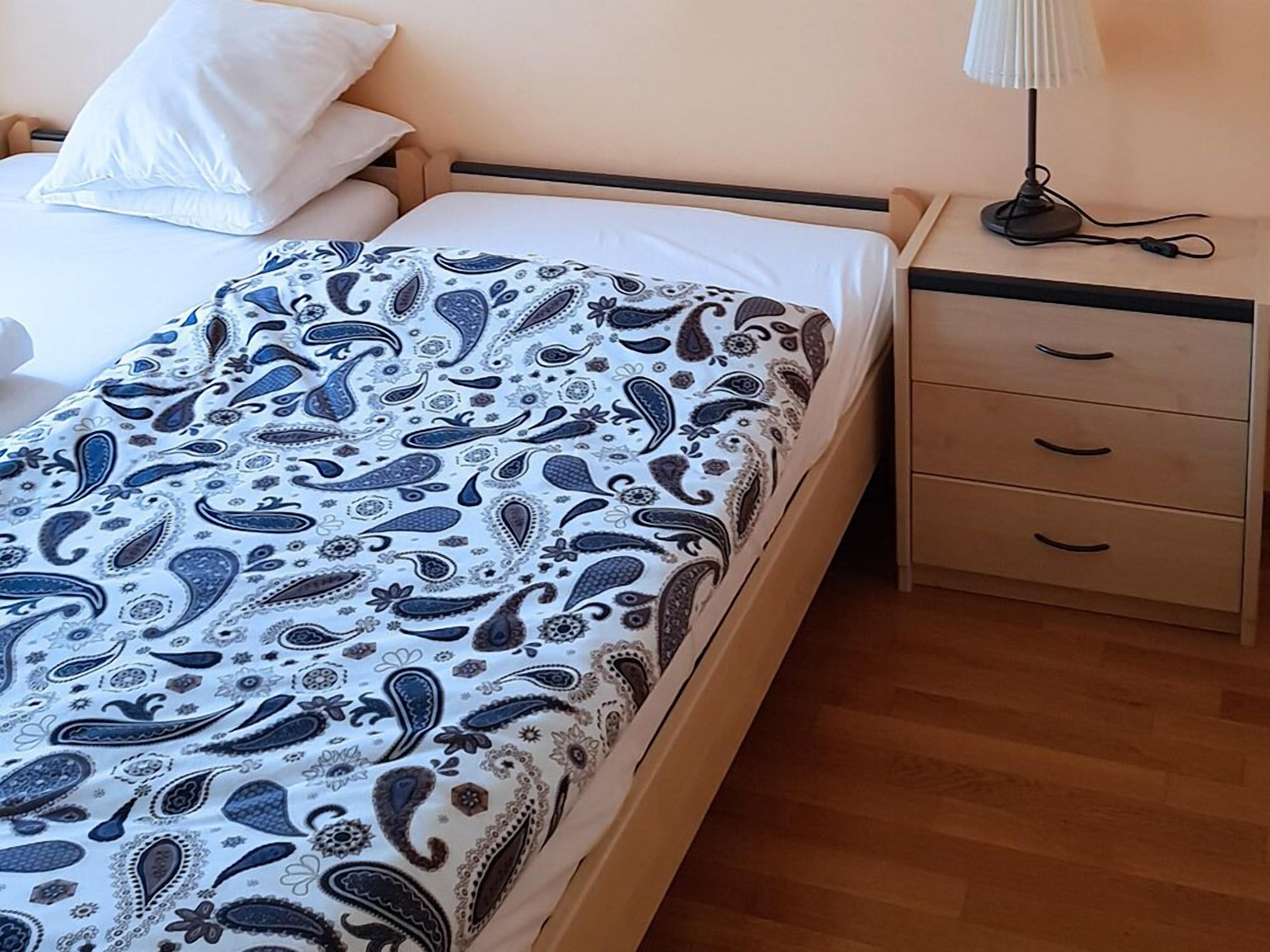 A bed with a patterned bedspread, a bedside table with a lamp, and a drawer unit.