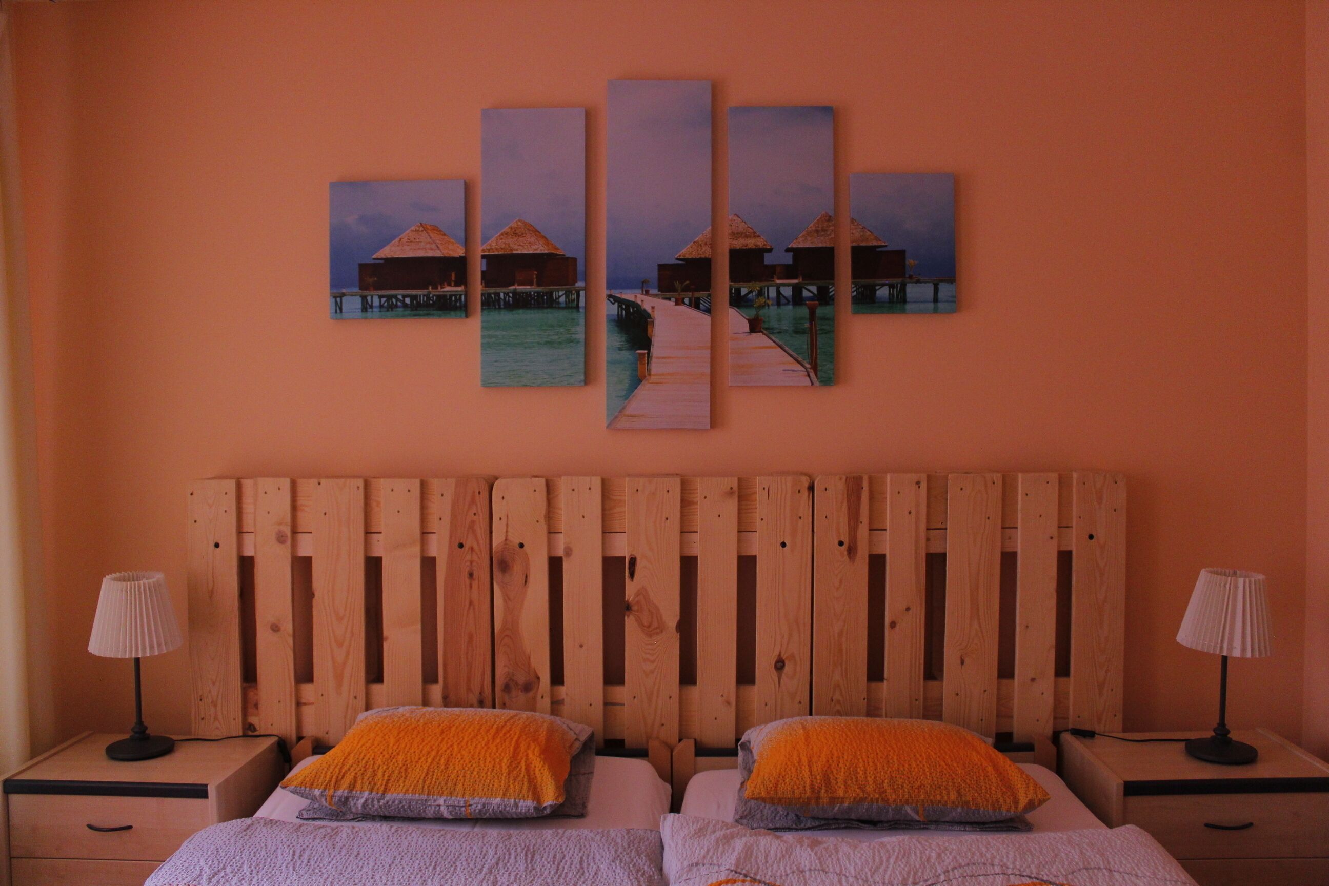 A bedroom with a wooden headboard, two beds with orange and grey bedding, two bedside tables with lamps, and a wall-mounted artwork of overwater bungalows.