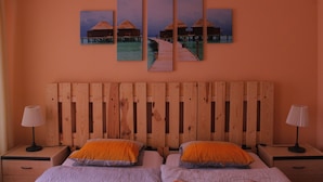 Quarto com cabeceira de madeira, duas camas com almofadas laranja e cinzentas, mesa de cabeceira de madeira com abajur e uma obra de arte na parede retratando bangalôs sobre a água.
