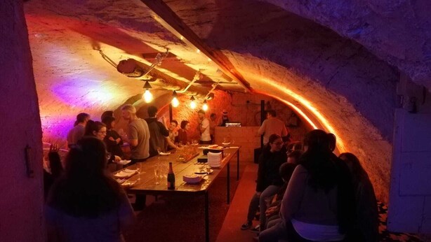 Uma área de refeições subterrânea com pouca luz, paredes de pedra e uma longa mesa posta para uma refeição.