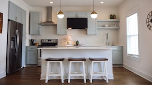 A modern kitchen with a central island, bar stools, stainless steel appliances, pendant lighting, and a large window. - Home on the Range (Cookeville)