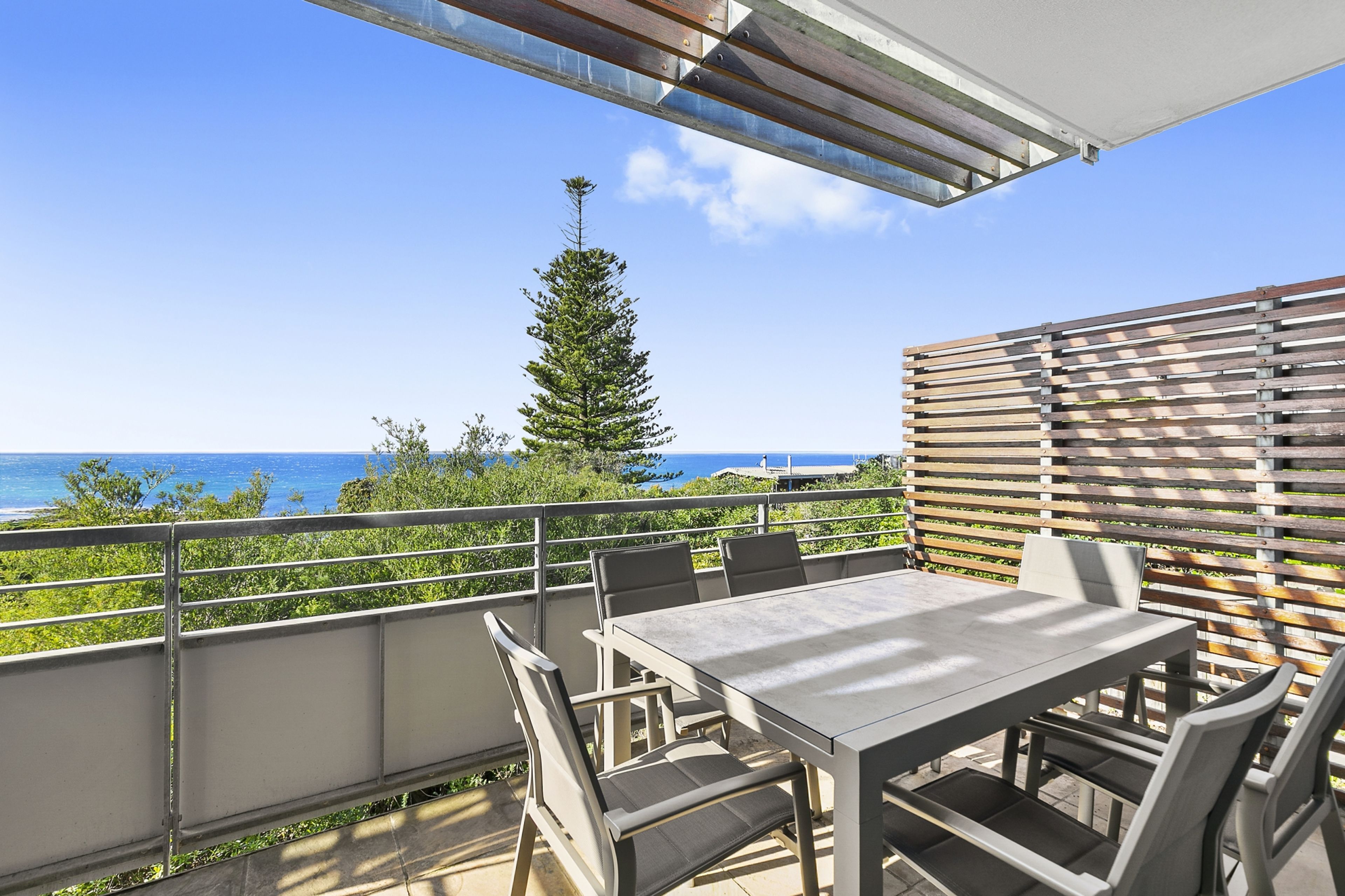 An outdoor patio with a table and chairs, overlooking a coastal view with a tree and a building in the distance.