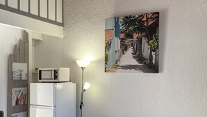 Een gezellige woonruimte met een grijze bank, een witte koelkast, een magnetron en een schilderij van een straatbeeld.