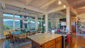 A spacious kitchen with large windows, a stone fireplace, and a dining area with a view of the mountains. - Sky High at Laurel Ridge | Post & Beam Cabin With Long-range Mountain Views! (Black Mountain)