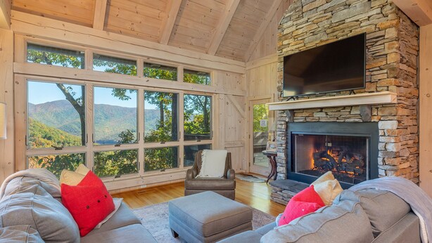 A cozy living room with a stone fireplace, large windows offering a mountain view, and comfortable seating. - Sky High at Laurel Ridge | Post & Beam Cabin With Long-range Mountain Views! (Black Mountain)