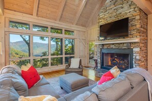 A cozy living room with a stone fireplace, large windows offering a mountain view, and comfortable seating.