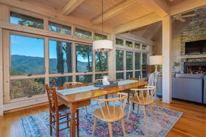 A spacious dining area with a wooden table and chairs, large windows offering a view of the mountains, and a stone fireplace.