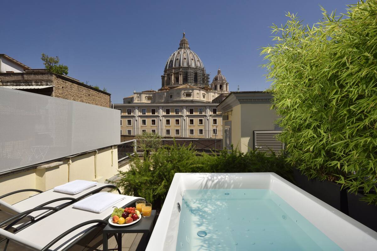 A rooftop terrace with a hot tub, lounge chairs, and a table with fruit and drinks. In the background, there is a large building with a dome.