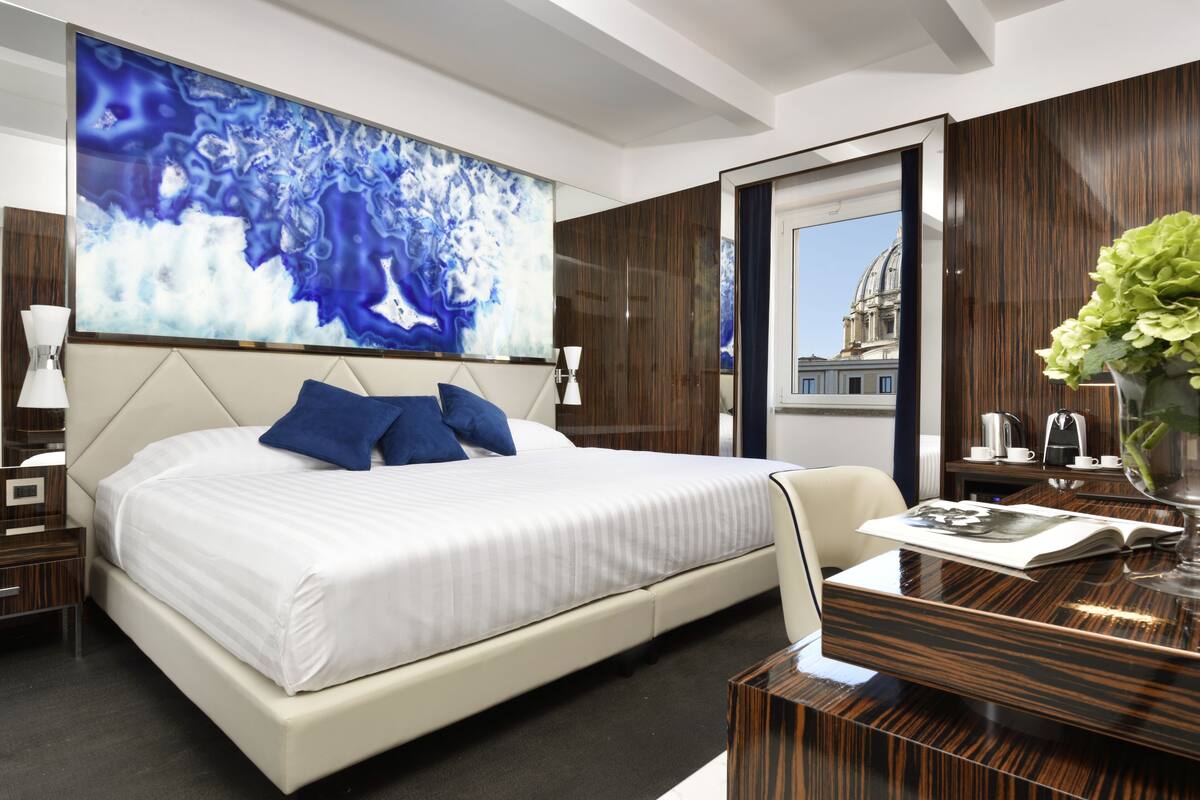 A modern hotel room with a large bed, a desk with a vase, and a view of a building through a mirror.