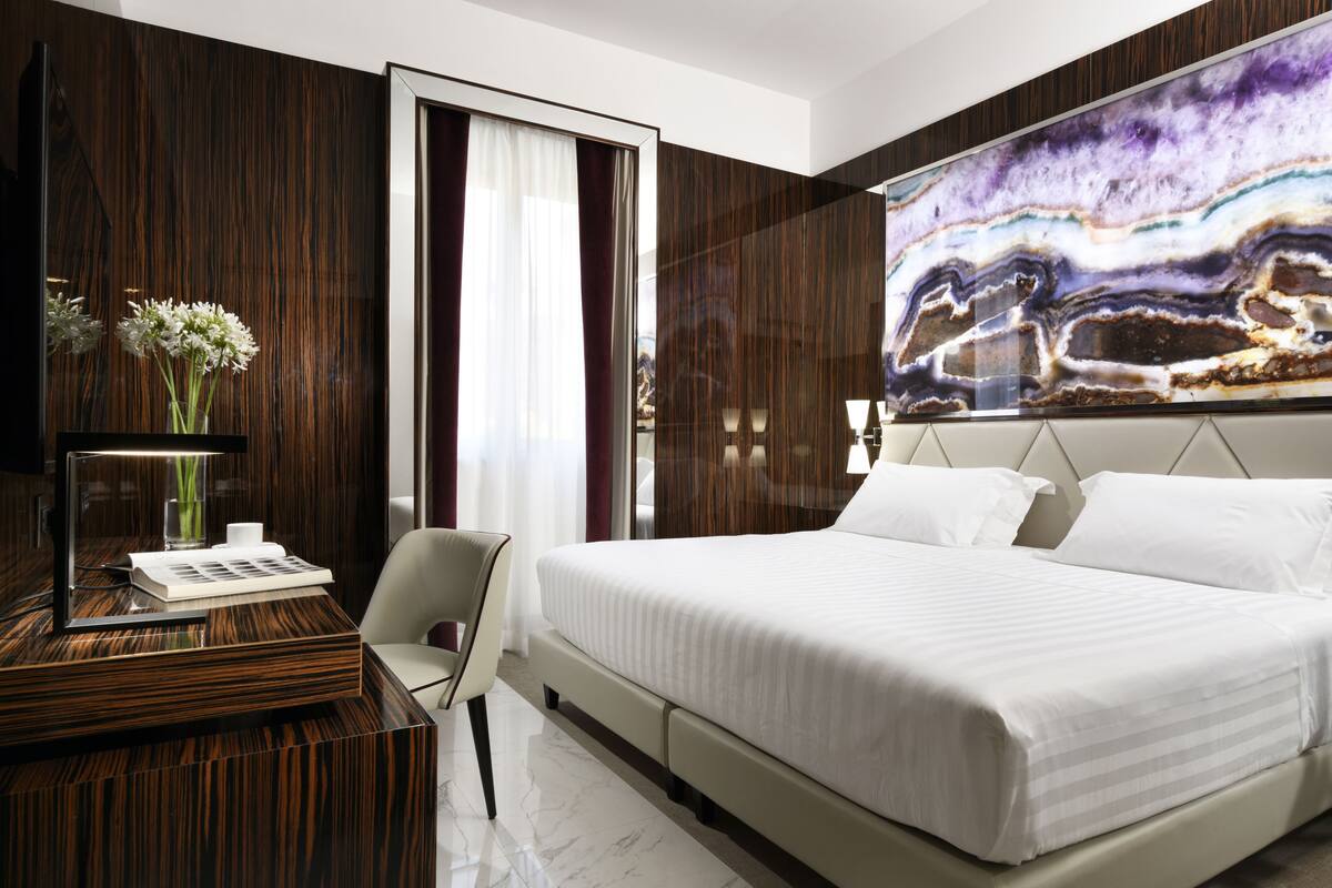 A modern hotel room with a large bed, a desk with a mirror, a vase with flowers, and a large abstract painting on the wall.