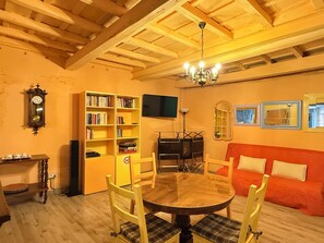 A room with a wooden ceiling, a round table with chairs, a bookshelf, a television, and a sofa.