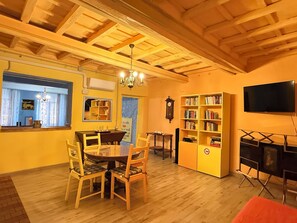 A dining area with a wooden table and chairs, a bookshelf, a television, and a clock on the wall.
