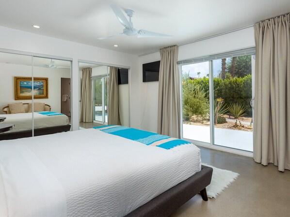 Un dormitorio con cama, ventilador de techo, ventana grande con cortinas y vistas a la vegetación.