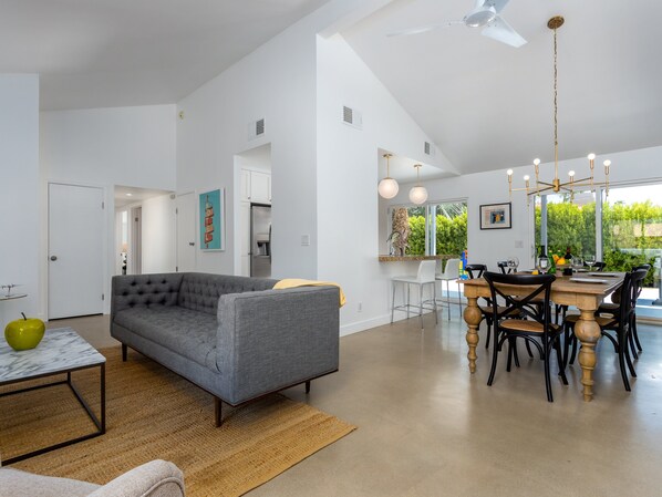 A modern living room with a grey sofa, a dining table with chairs, a kitchen area with a refrigerator, and a large window with a view of greenery. - Casa Bonita - Private Pool & Mountain Views (Palm Springs)