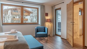 A hotel room with a bed, a chair, a lamp, and a large window with a view of buildings. - FINEST VILLA Zell am See, luxury, sauna, 12 pax (Zell am See)