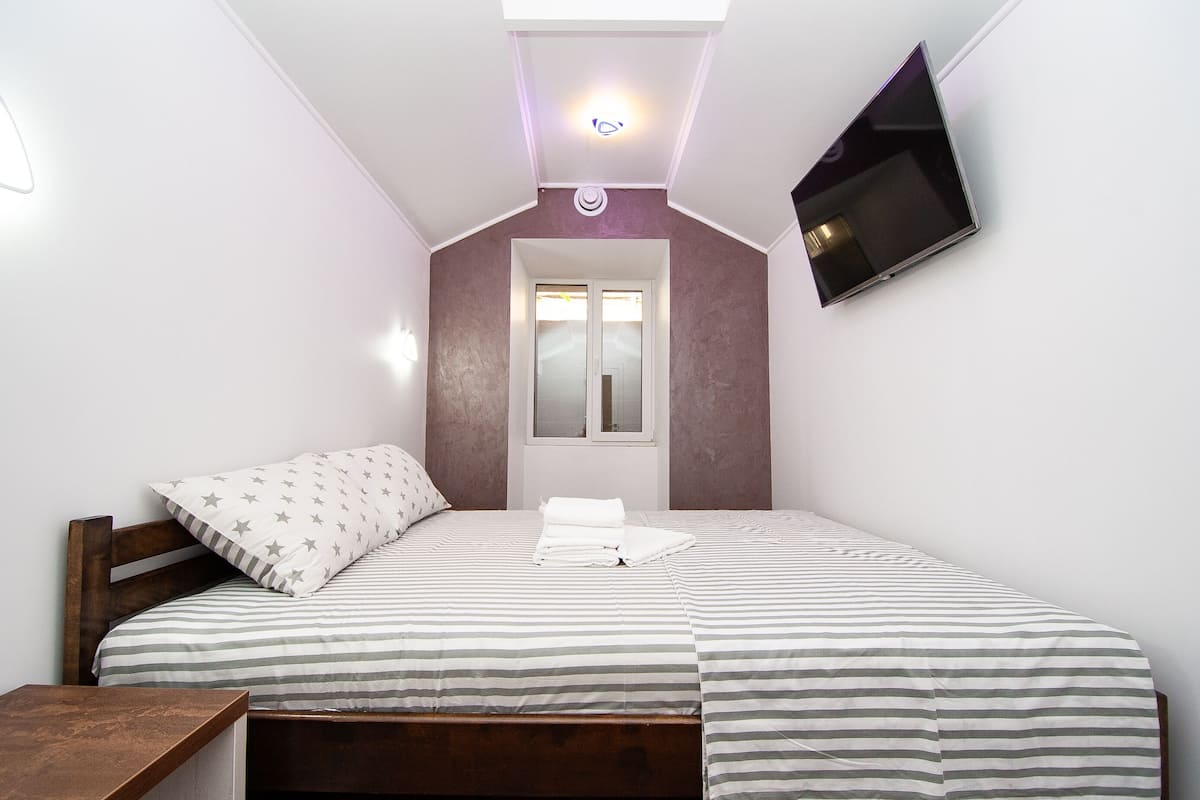 Un dormitorio con cama, cabecera de madera, mesita de noche, ventana y televisor fijado a la pared.