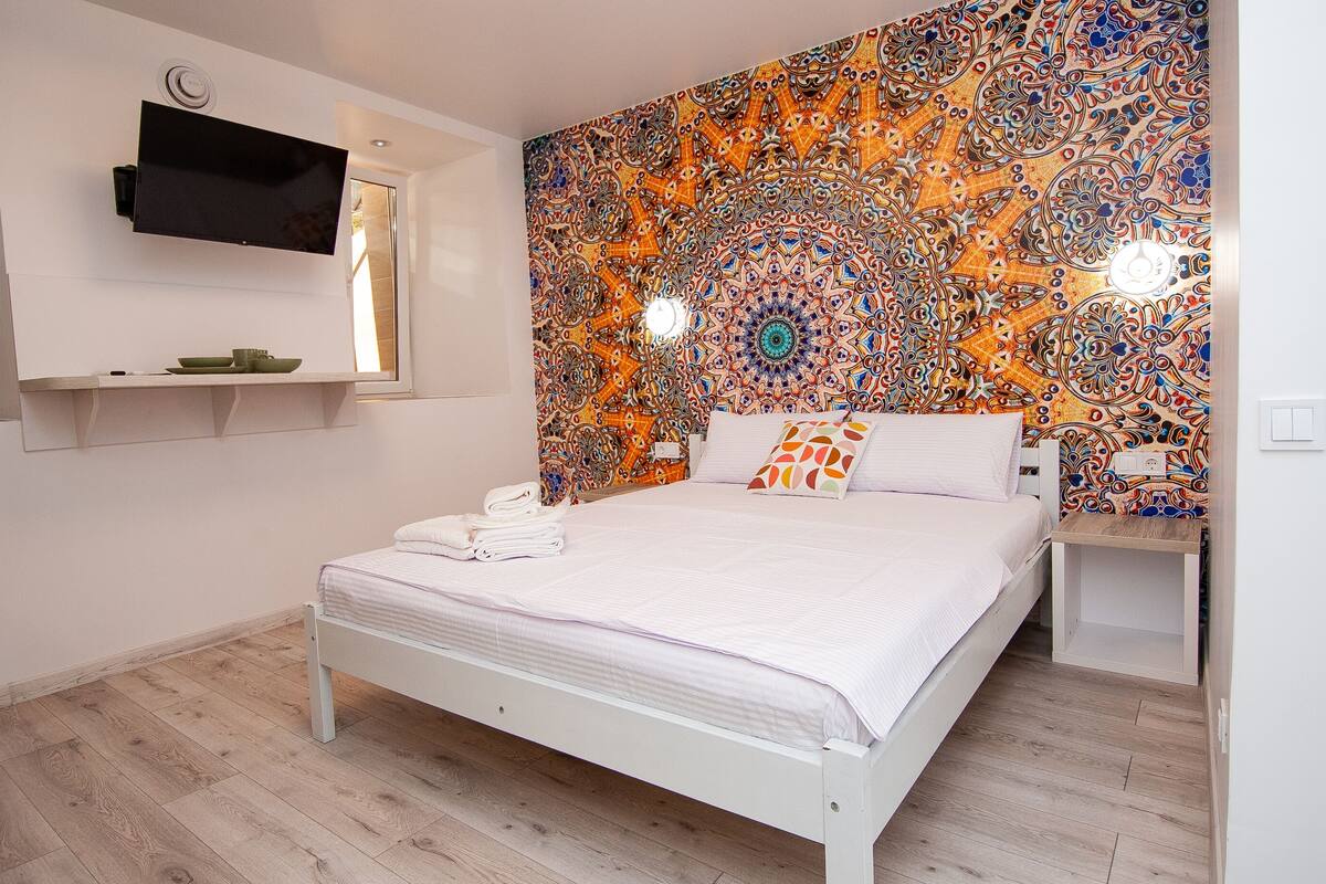 Un dormitorio con cama, televisor de pantalla plana montado en la pared y un tapiz colorido detrás de la cama.