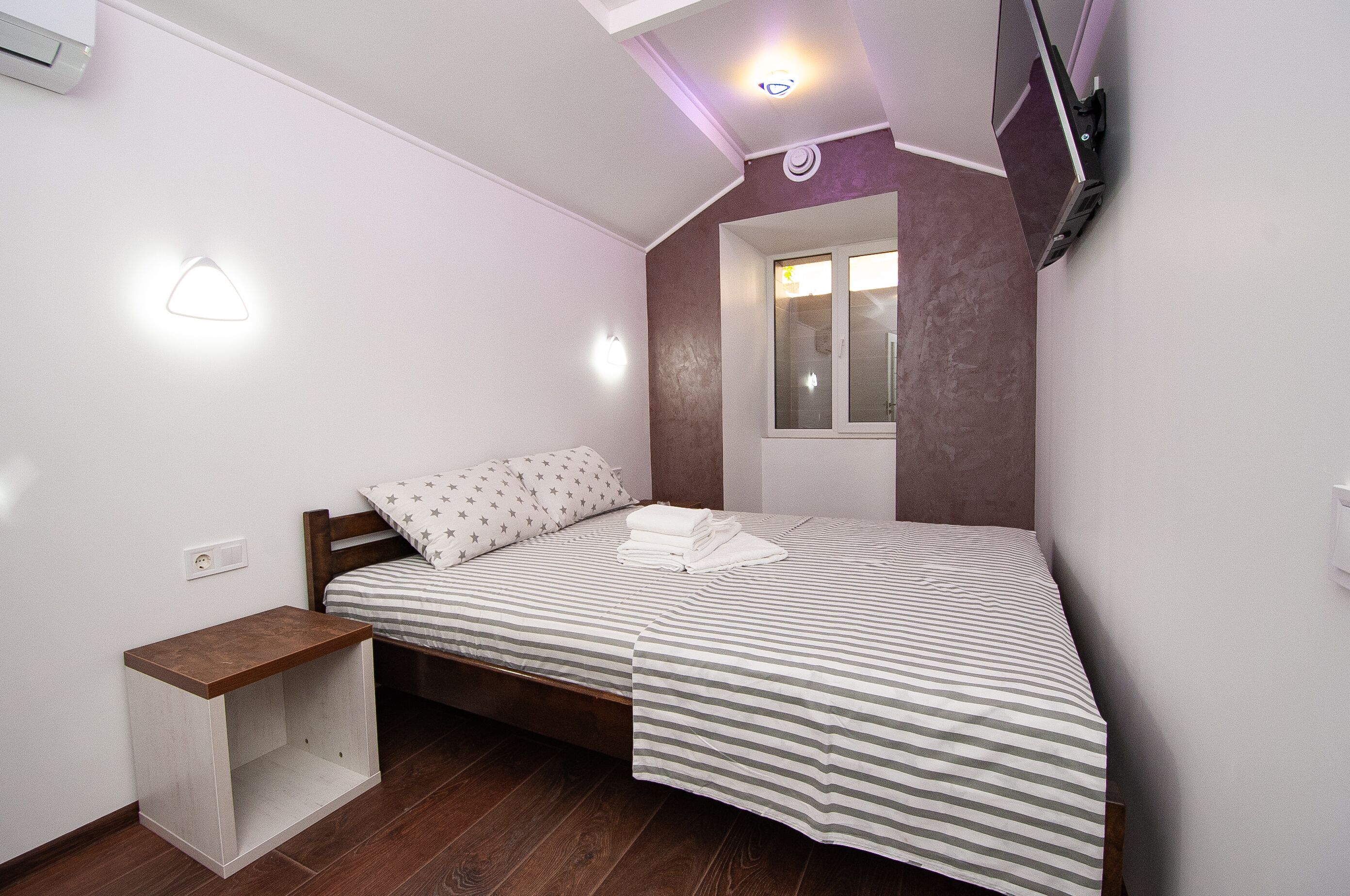 Uma cama bem arrumada com roupa de cama listrada, uma mesinha de cabeceira de madeira, uma televisão na parede e uma janela com vista para o exterior.