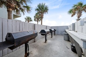 An outdoor barbecue area with multiple grills, a wooden fence, and palm trees.