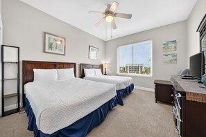 A hotel room with two beds, a ceiling fan, a desk with a computer, and a window with a view of buildings.