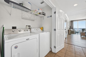 Ein Waschraum mit einer Maytag Waschmaschine und einem Trockner, ein Metallregal mit Kartons und Blick auf einen Balkon mit Gartenmöbeln.