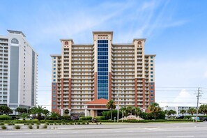 A multi-story hotel with a central glass facade, surrounded by greenery and a clear sky.