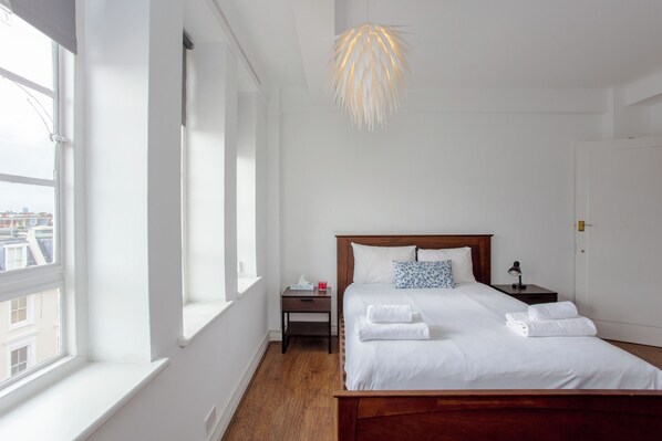 A hotel room with a large window, a wooden bed with white linens, a bedside table with a lamp, and a hanging light fixture.