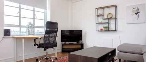 Una sala de estar moderna con un escritorio, una silla de oficina negra, un sofá gris, un televisor y un estante decorativo con un espejo.