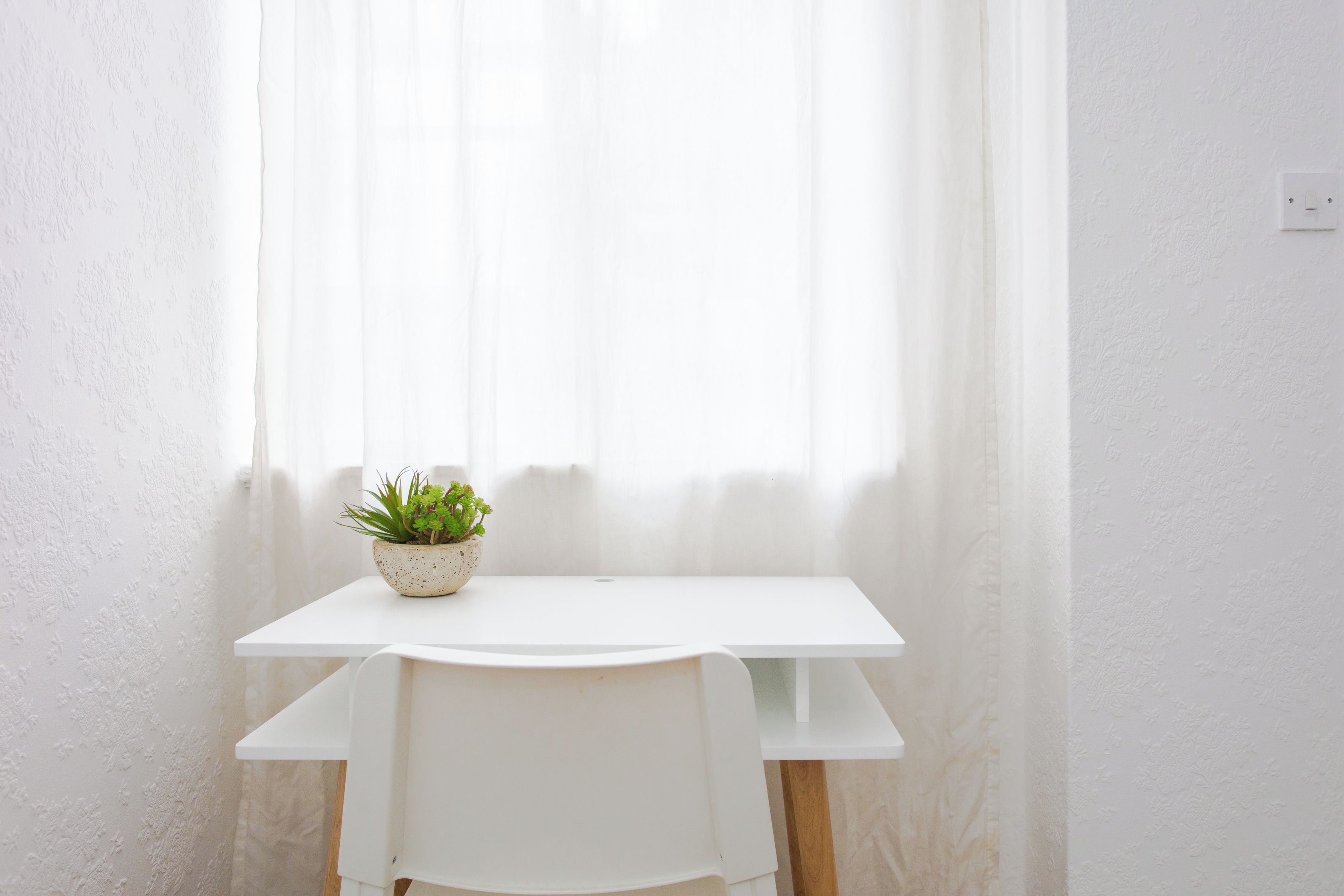 Una mesa blanca con una planta en maceta, una silla blanca y una ventana con cortinas translúcidas.
