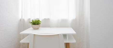 Una mesa blanca con una planta en maceta, una silla blanca y una ventana con cortinas translúcidas.
