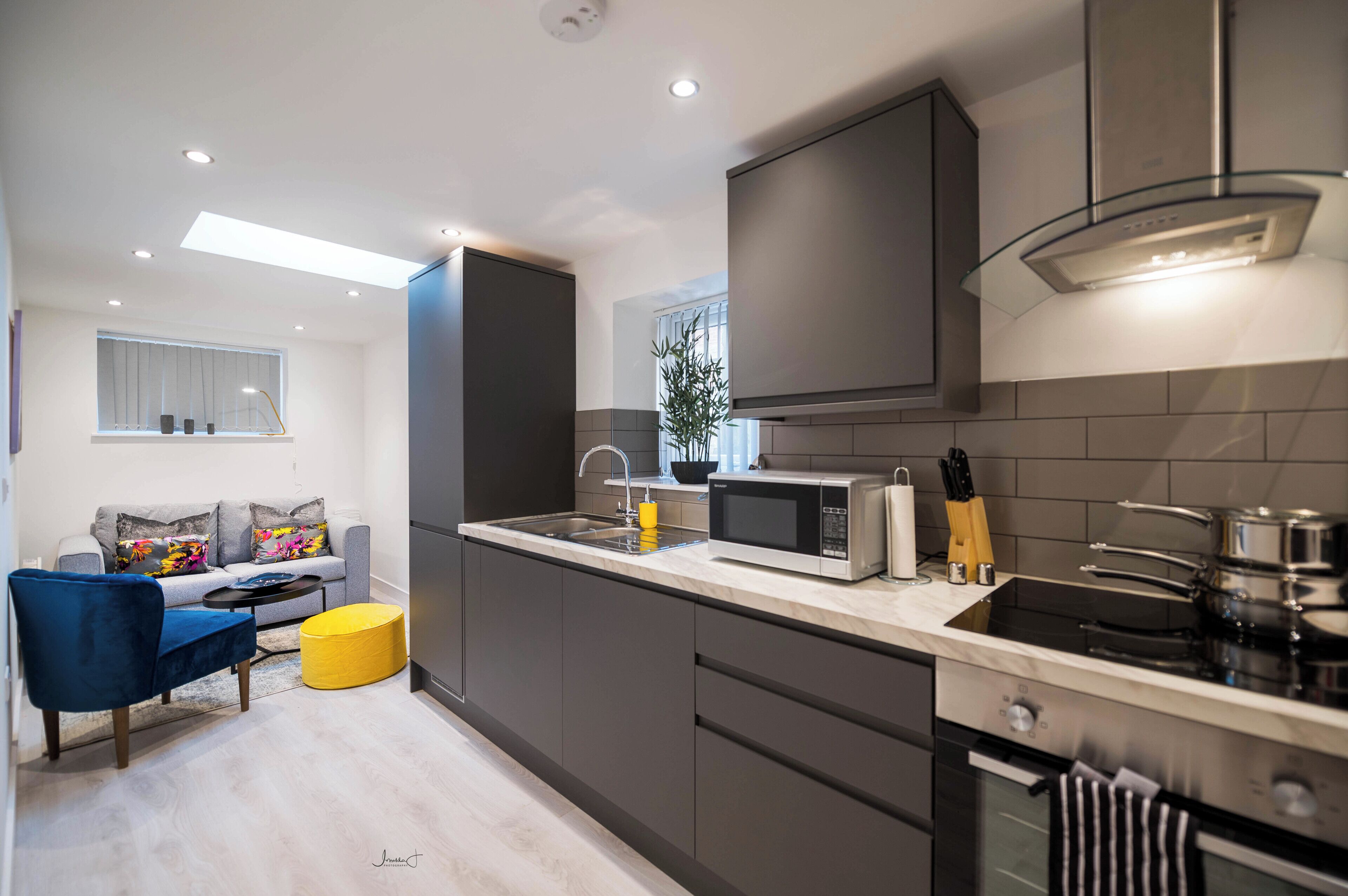 A modern kitchen with a built-in microwave, stove, and sink. There is a dining area with a blue chair and a yellow ottoman.